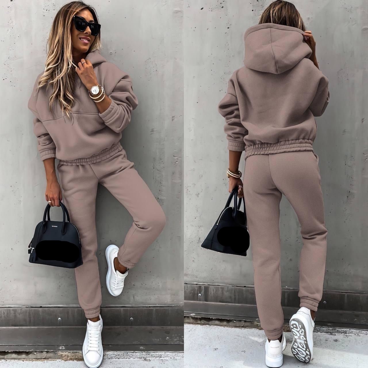Een vrouw draagt een lichtbruin joggingpak, bestaande uit een oversized hoodie en een bijpassende broek met elastische tailleband. Ze combineert het met witte sneakers en een zwart handtasje van Balenciaga. Haar stijl wordt compleet gemaakt met zonnebrillen en een elegant horloge. De comfortabele en stijlvolle outfit is perfect voor een casual, modieuze look.