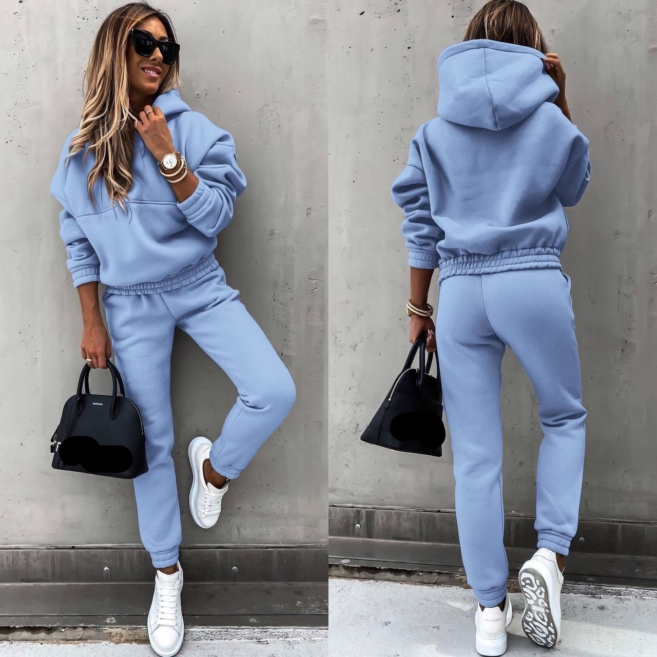 Een vrouw draagt een lichtblauw joggingpak, bestaande uit een oversized hoodie en een bijpassende broek met een elastische tailleband. Ze draagt witte sneakers en een zwart handtasje van Balenciaga. Haar look wordt afgemaakt met zonnebrillen en een elegant horloge. Deze comfortabele en stijlvolle outfit is perfect voor een casual, modieuze uitstraling.