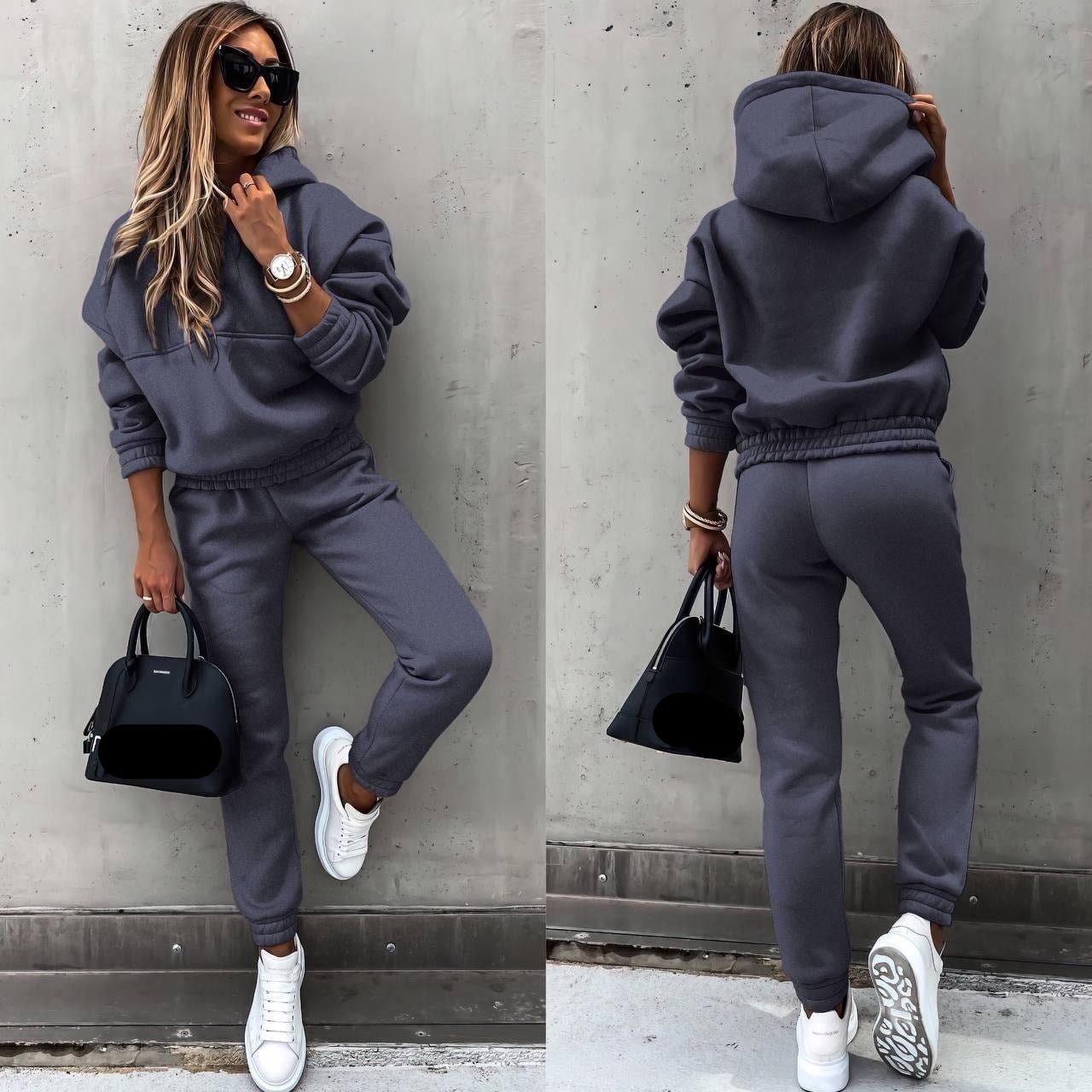 Een vrouw draagt een donkergrijs joggingpak, bestaande uit een oversized hoodie en een bijpassende broek met een elastische tailleband. Ze heeft witte sneakers aan en draagt een zwart handtasje van Balenciaga. Haar look wordt afgemaakt met zonnebrillen en een elegant horloge, wat haar casual en stijlvolle uitstraling versterkt. Perfect voor comfortabele, modieuze outfits.