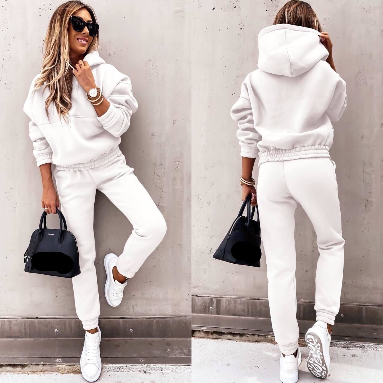 Een vrouw in een elegante, witte jogging-set met een oversized hoodie en bijpassende broek. Ze draagt witte sneakers en een zwart Balenciaga handtasje. De look wordt compleet met een horloge en zonnebrillen, wat haar stijlvolle, casual uitstraling benadrukt. Perfect voor een comfortabele en modieuze outfit.