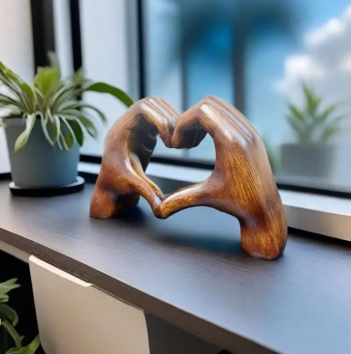Een betoverend houten hartbeeld van Olivan, met twee handen die samen een hart vormen. Het kunstwerk staat op een boekenplank, omringd door planten, en voegt een elegante, warme sfeer toe aan het interieur.
