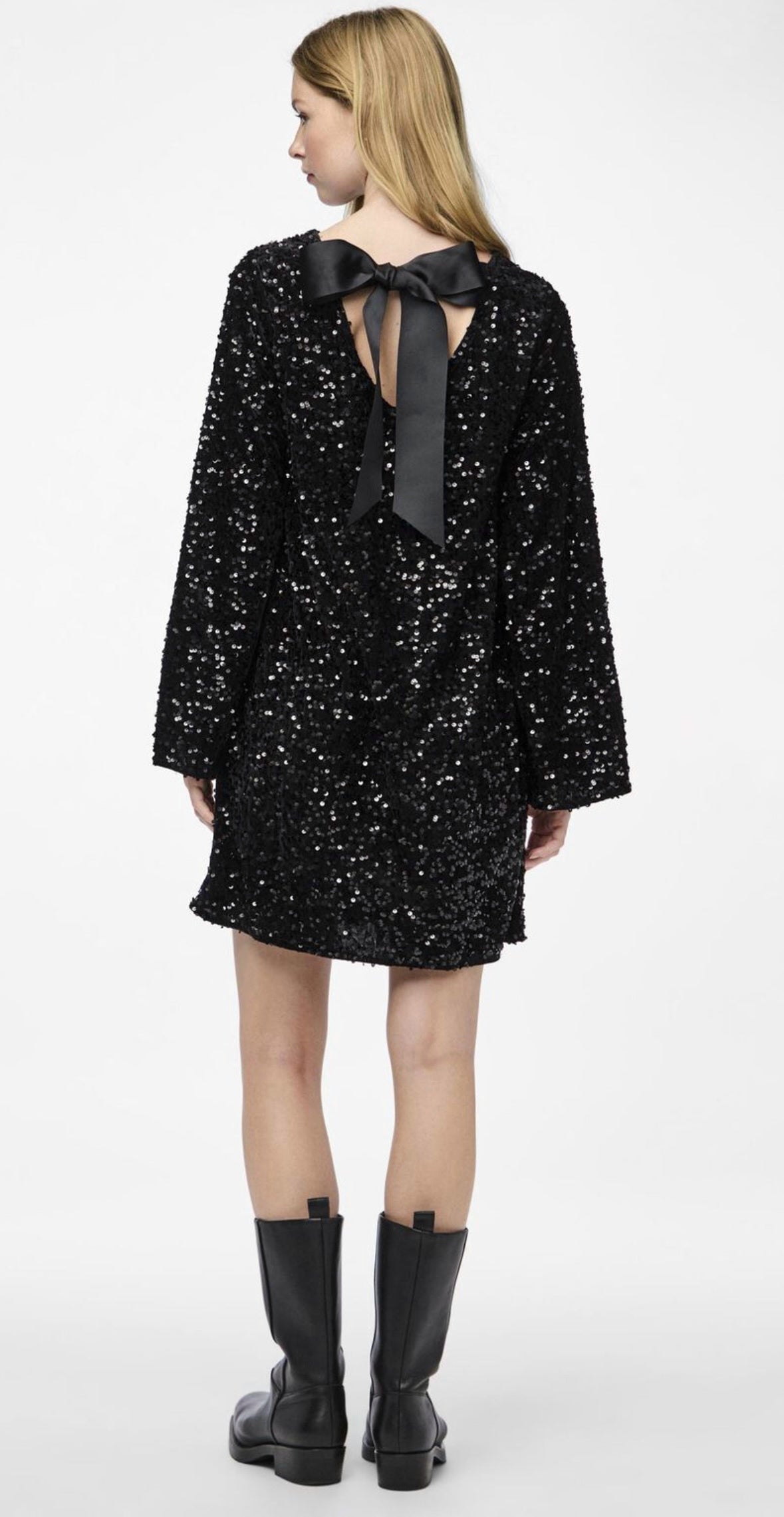 Model draagt de Ophira Dreamwear Elsa Glitter Jurk in zwart, versierd met glinsterende pailletten en een elegante zwarte strik aan de achterkant, gecombineerd met zwarte laarzen.