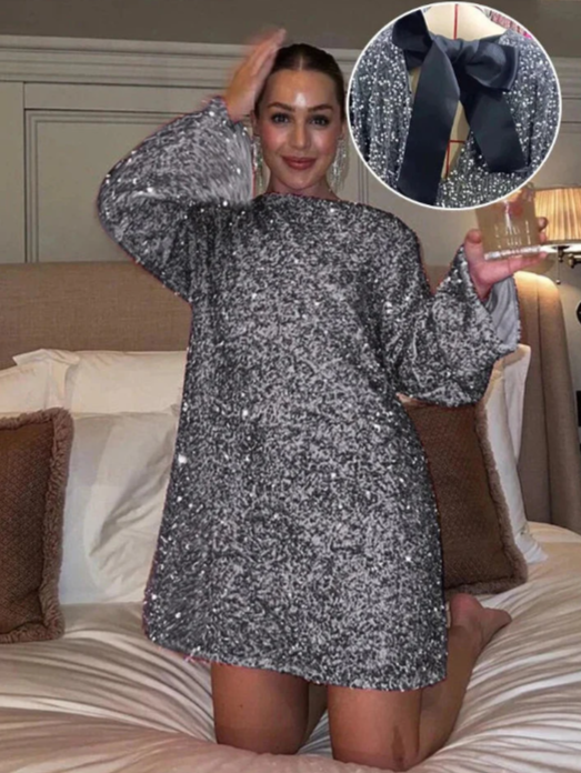 Model draagt de Ophira Dreamwear Elsa Glitter Jurk in zilver met glinsterende pailletten, terwijl ze in een slaapkamer poseert met een glazen drankje in haar hand en haar haren lichtjes aanraakt, met de zwart satin strik op de achterkant zichtbaar in de afbeelding.
