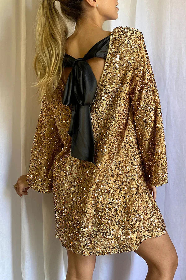 Model draagt de Ophira Dreamwear Elsa Glitter Jurk in goud met glinsterende pailletten en een grote zwarte strik op de achterkant, terwijl ze poseert met haar haar in een hoge staart.