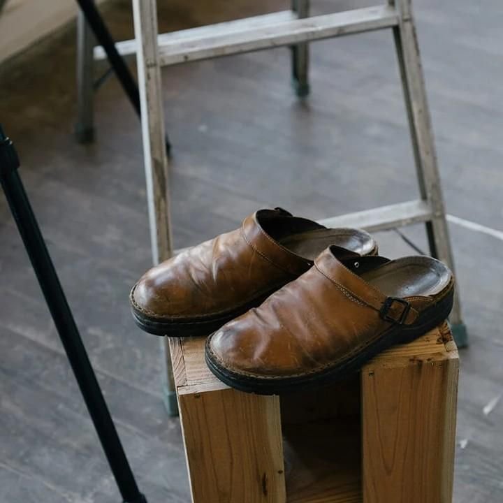 Bruine leren orthopedische slippers op een houten blok, met een ladder op de achtergrond.