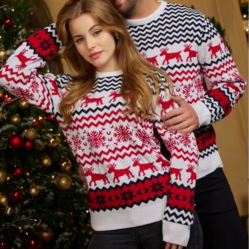Een man en een vrouw dragen bijpassende kersttruien met een wit, rood en zwart patroon van sneeuwvlokken, zigzag lijnen en rendieren. De truien zijn stijlvol en comfortabel, perfect voor de feestdagen. Ze staan voor een kerstboom, wat de feestelijke sfeer benadrukt.