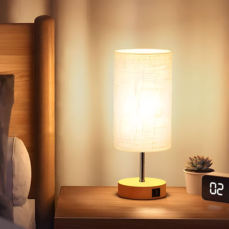 Solvani bedlamp met een warme en moderne uitstraling, geplaatst op een nachtkastje naast een bed. De lamp heeft een cilindervormige kap en een houten voet met een ingebouwde USB-aansluiting, ideaal voor het opladen van apparaten. Perfect voor een sfeervolle en functionele toevoeging aan de slaapkamer.