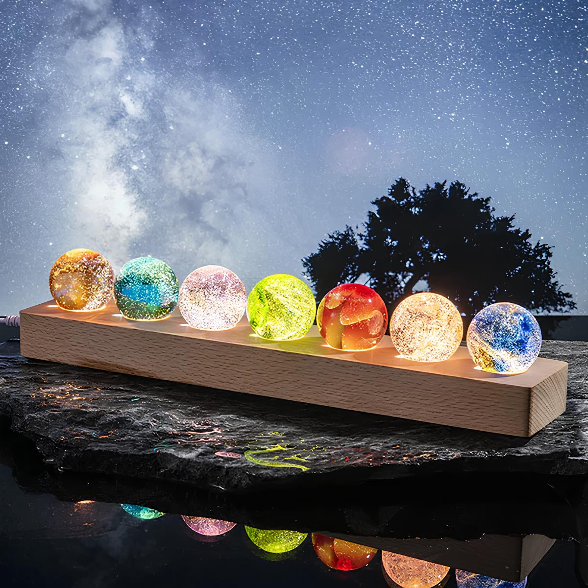 StarGlow kristallen lampen in een rij van kleurrijke bollen op een houten basis, verlicht tegen een betoverende sterrenhemel, die een magische en rustgevende sfeer creëren.