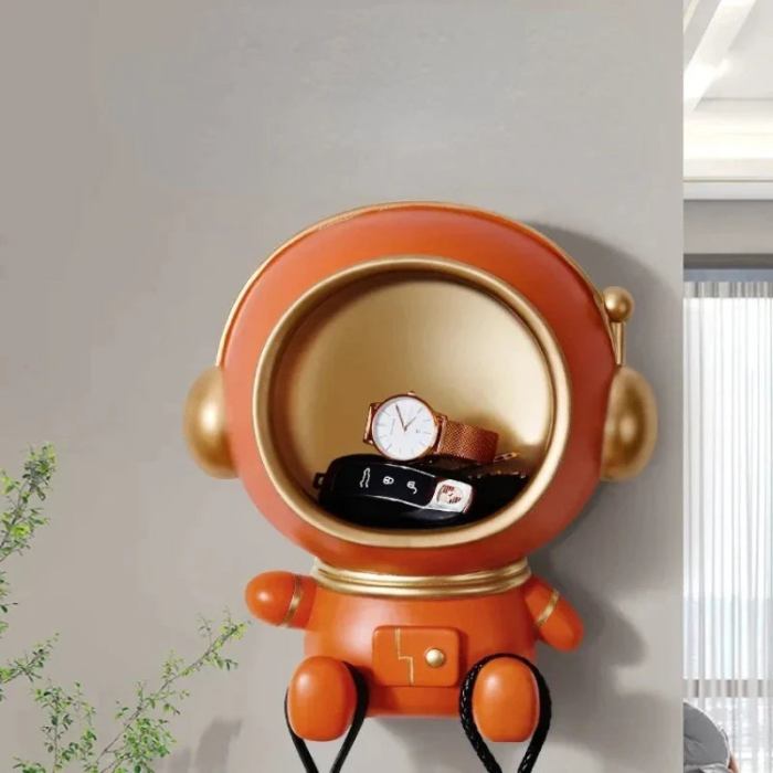 Een astronaut-sleutelhanger in oranje met gouden details, bevestigd aan de muur en ontworpen om kleine voorwerpen op te bergen. Het open vak van de astronaut bevat een horloge en sleutels. De speelse en functionele opberger heeft ook een haak voor het ophangen van tassen of andere accessoires, ideaal voor het organiseren van spullen in een modern interieur.