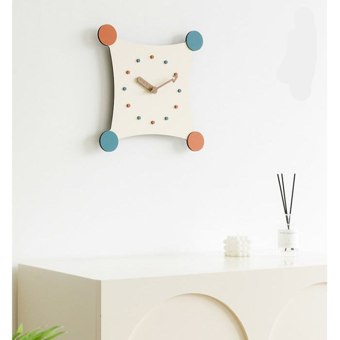 Een moderne wandklok met een speelse uitstraling, bevestigd aan een witte muur. De klok heeft een vierkante vorm met afgeronde hoeken en kleurrijke stippen in oranje, groen en blauw als urenmarkeringen. De houten wijzers passen perfect bij het minimalistische ontwerp, waardoor het een eigentijdse toevoeging is aan het interieur. De klok staat op een kast met enkele decoratieve items ernaast.
