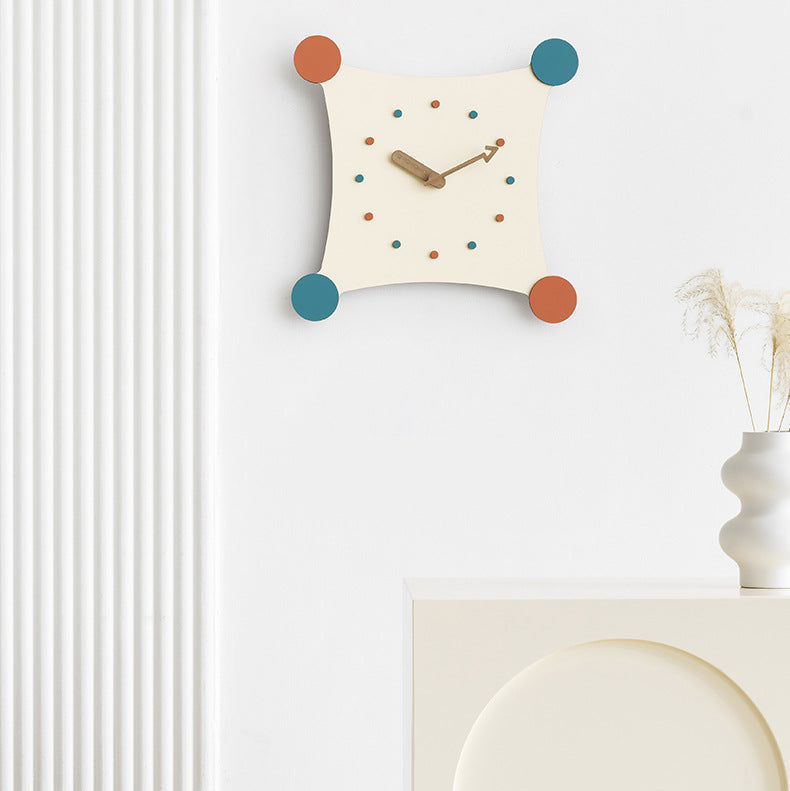 Een moderne wandklok met een vierkante, zachte vorm, bevestigd aan een witte muur. De klok heeft kleurrijke stippen in oranje, blauw en groen op de hoeken, met houten wijzers en een minimalistisch ontwerp. Onder de klok staat een eenvoudige kast met een elegante vaas met droogbloemen. Het ontwerp is strak en eigentijds, waardoor het een perfecte aanvulling is voor moderne interieurs.