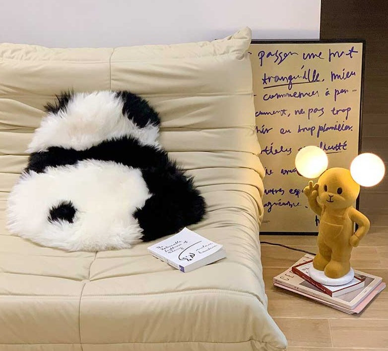 Een zwart-wit panda-vormig vloerkleed ligt op een beige bank, naast een stapel boeken en een schattige lamp in de vorm van een geel teddybeer. Het zachte, harige kleed biedt bescherming aan de bank en voegt een speelse, gezellige sfeer toe aan het interieur. Het unieke ontwerp van het vloerkleed maakt het een charmant accessoire in de woonkamer.