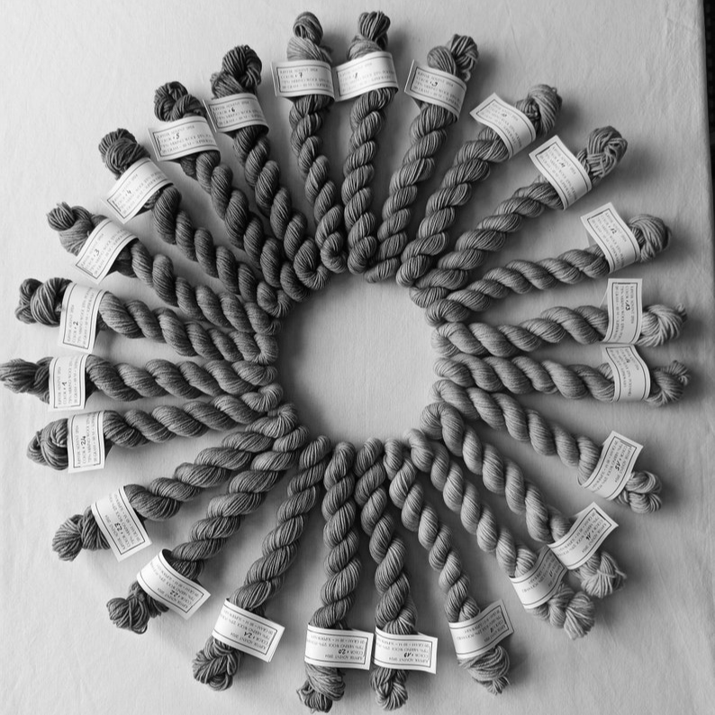 Een cirkel van zorgvuldig gerangschikte garenstrengen, elk met een label, die een prachtig patroon vormen. Deze afbeelding toont de variëteit en kleuren van garen die te vinden zijn in een adventskalender. Het biedt een inspirerende en betoverende ervaring voor handwerkers die op zoek zijn naar nieuwe kleuren om hun projecten mee te verrijken, met elke streng een verrassend tintje voor iedere dag.