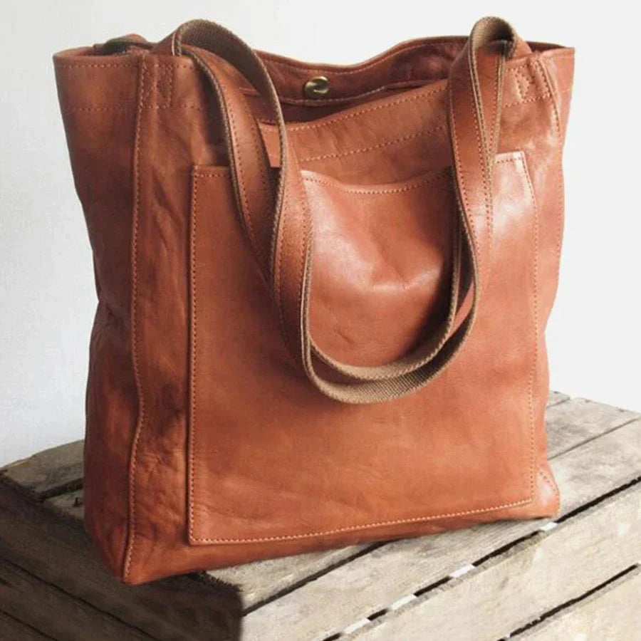 Een elegante leren schoudertas in bruin, met een groot, plat ontwerp en stevige handvatten. De tas heeft een voorzak voor extra opbergruimte en is ideaal voor dagelijks gebruik of als stijlvolle werk- en winkel tas.