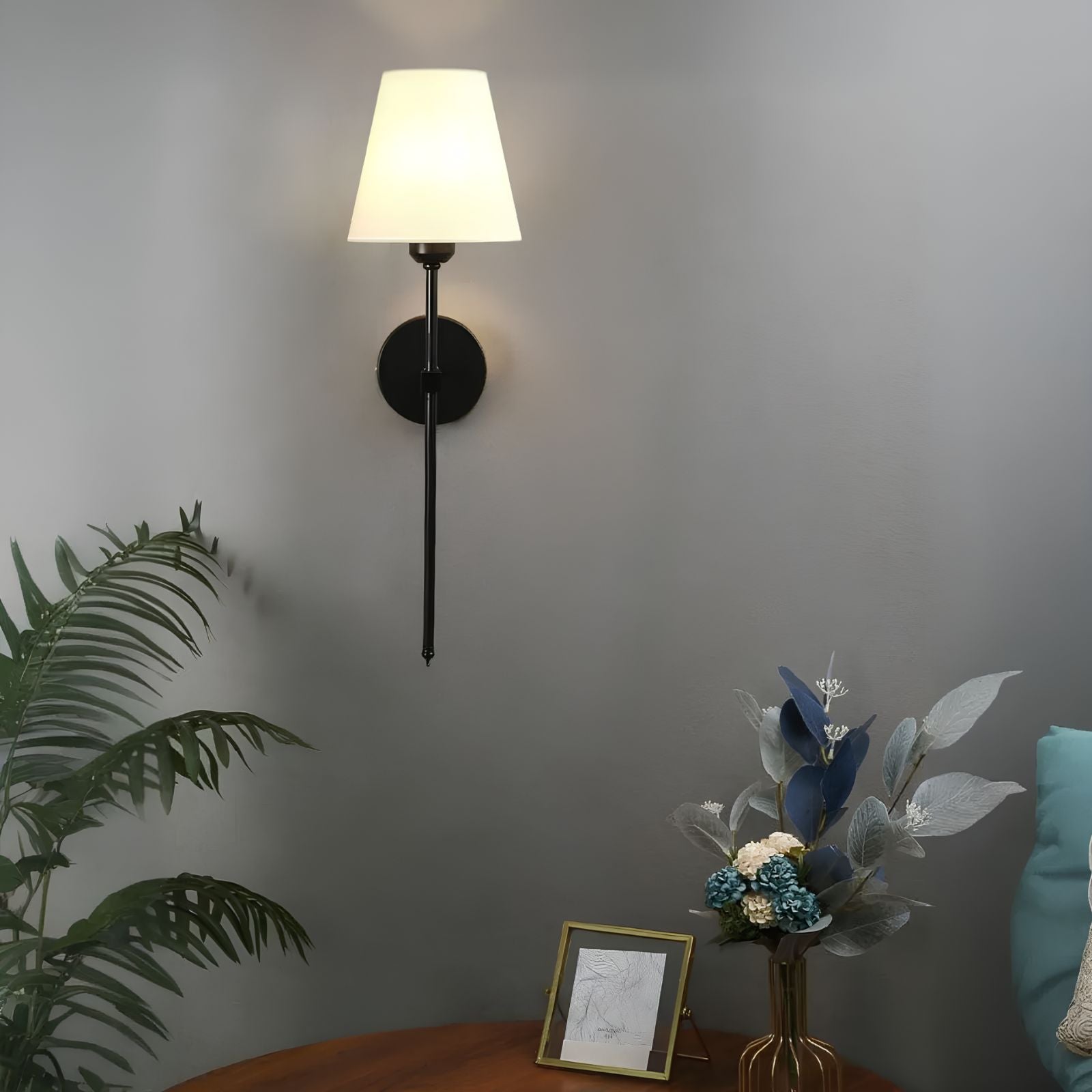 De Aurelia Wall Light met een klassieke zwarte afwerking en een witte lampenkap verlicht een stijlvolle ruimte. De wandlamp is bevestigd aan een grijze muur en verfraaid met een plant, bloemen en een ingelijste tekening op een houten tafel, waardoor een warme en verfijnde sfeer ontstaat.
