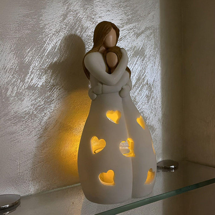 Tijdloze kandelaar in de vorm van een moeder die haar kind omhelst, met hartvormige uitsparingen die warm licht verspreiden. De kandelaar staat op een glazen plank en werpt een sfeervolle gloed op de muur. Perfect als decoratief element of als betekenisvol cadeau.