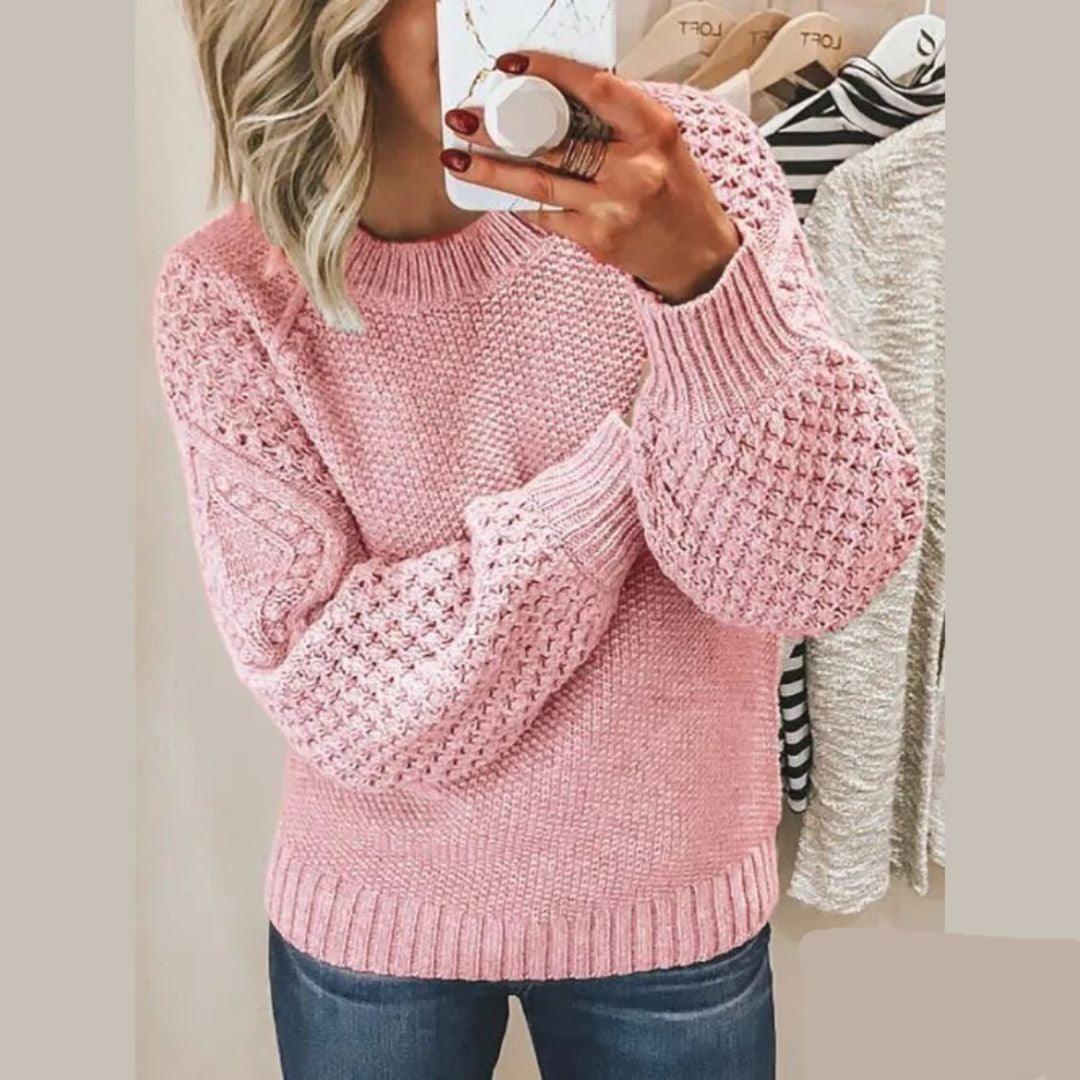 Een vrouw draagt een zachte, gebreide trui in lichtroze met een opvallend textuurpatroon op de mouwen en schouders. Ze houdt een kop koffie vast en kijkt in de spiegel. De trui heeft een ronde hals en biedt zowel comfort als stijl, perfect voor een casual, modieuze uitstraling.