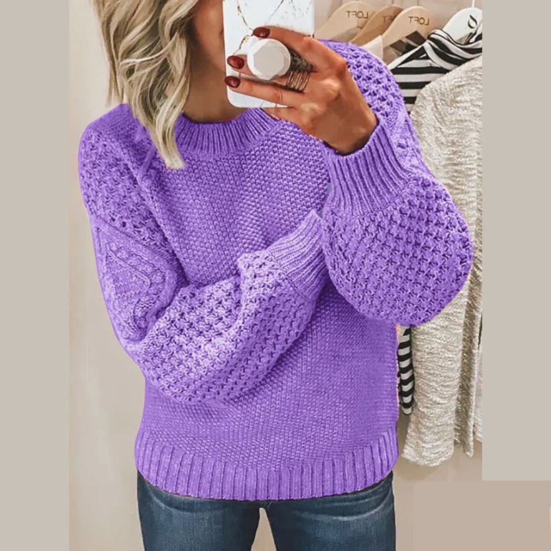 Een vrouw draagt een zachte, gebreide trui in lila met een opvallend textuurpatroon op de mouwen en schouders. Ze houdt een kop koffie vast en kijkt in de spiegel. De trui heeft een ronde hals en biedt zowel comfort als stijl, ideaal voor een casual, modieuze uitstraling.