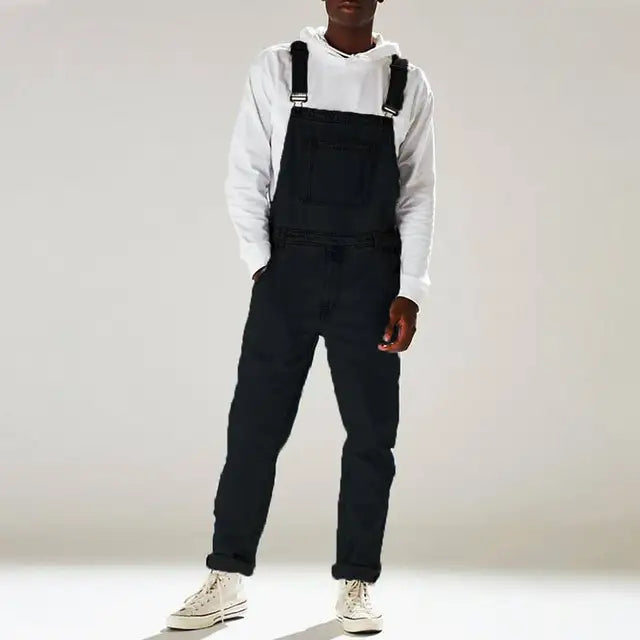 Man draagt een donkerblauwe denim jumpsuit met een moderne en casual uitstraling. Het kledingstuk heeft een relaxte pasvorm, verstelbare schouderbanden en een zak aan de voorkant, gecombineerd met een witte hoodie en sneakers voor een trendy en comfortabele zomerse outfit. Perfect voor een casual, veelzijdige stijl.