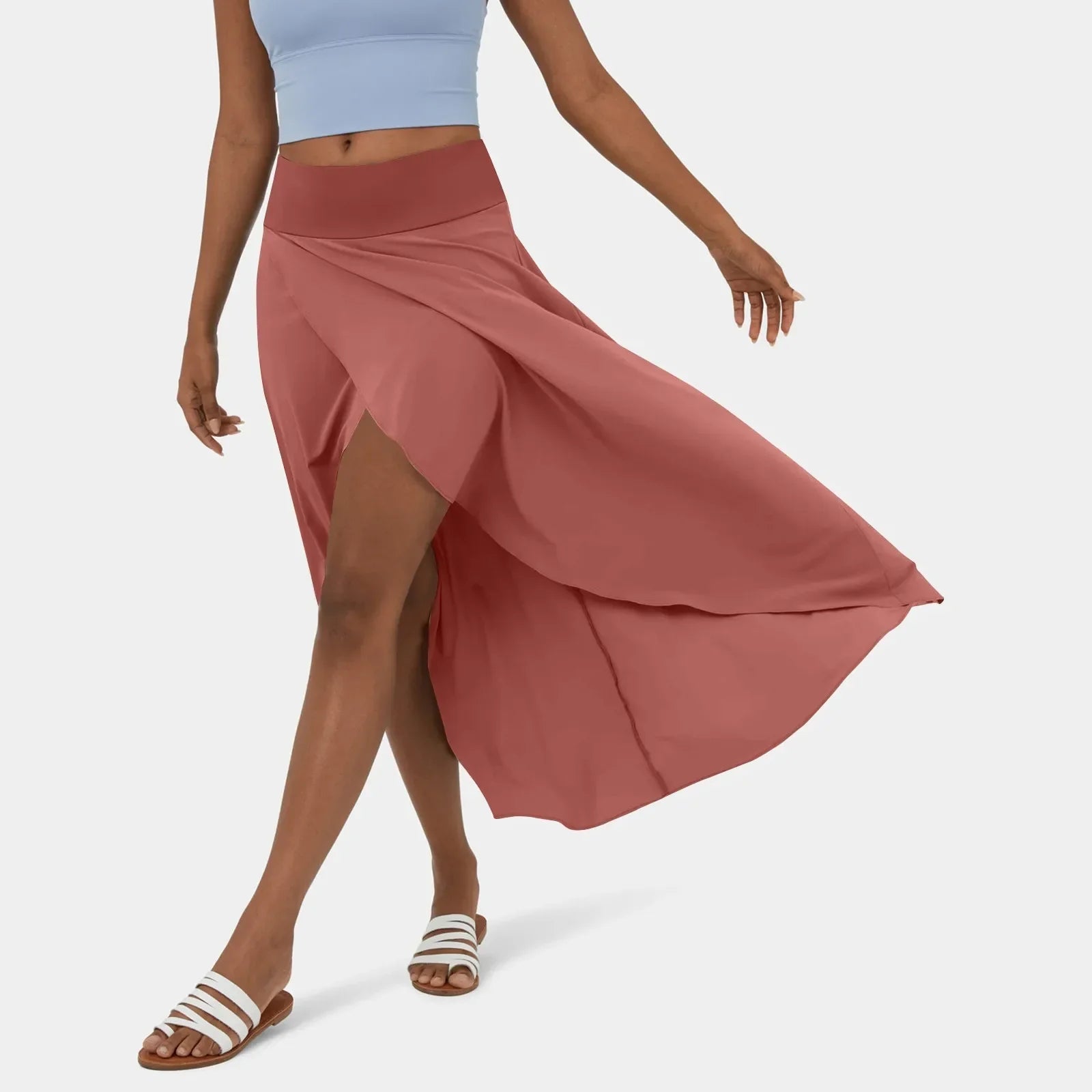 Een vrouw draagt een stijlvolle roze rok die sierlijk meebeweegt terwijl ze loopt. De rok heeft een vloeiende snit en een hoge taille, gecombineerd met een lichtblauw sporttopje en witte sandalen, perfect voor een casual en elegante zomerse uitstraling.