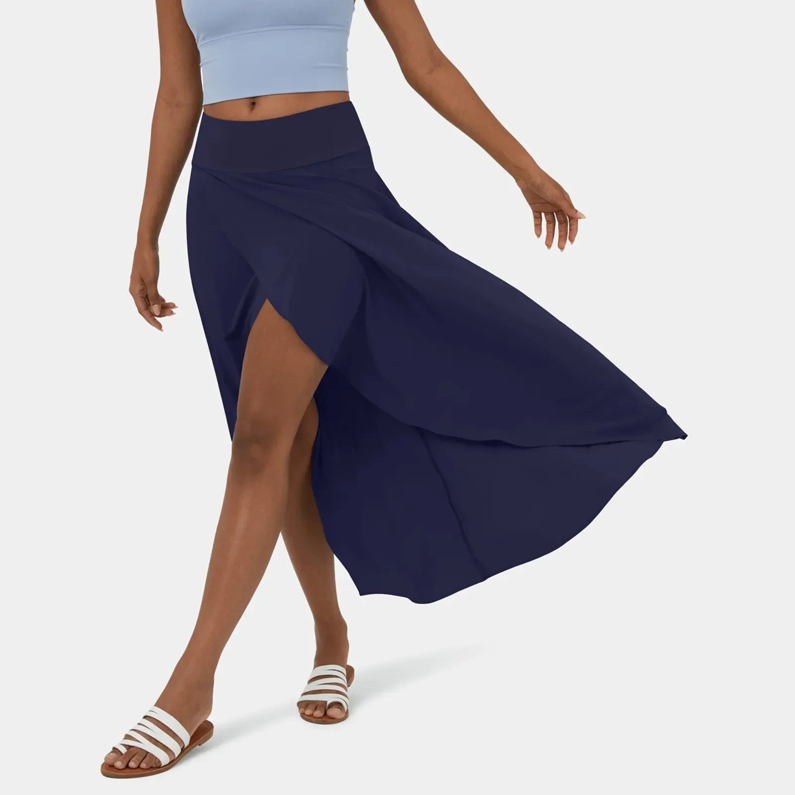 Een vrouw in een stijlvolle groenblauwe rok die sierlijk meebeweegt terwijl ze loopt. De rok heeft een elegante snit met een hoge taille en is gecombineerd met een lichtblauw sporttopje en witte sandalen, wat zorgt voor een frisse, zomerse uitstraling.