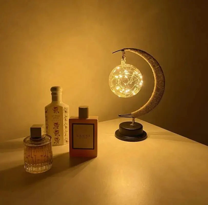 Een betoverende maanlamp die een magische gloed verspreidt, verlicht door delicate lichtjes in een gloeiende bol. De lamp staat op een elegante zwarte basis en wordt omringd door luxe parfumflessen, wat een luxe en serene sfeer creëert. Perfect voor een stijlvolle en warme ambiance in elke ruimte.