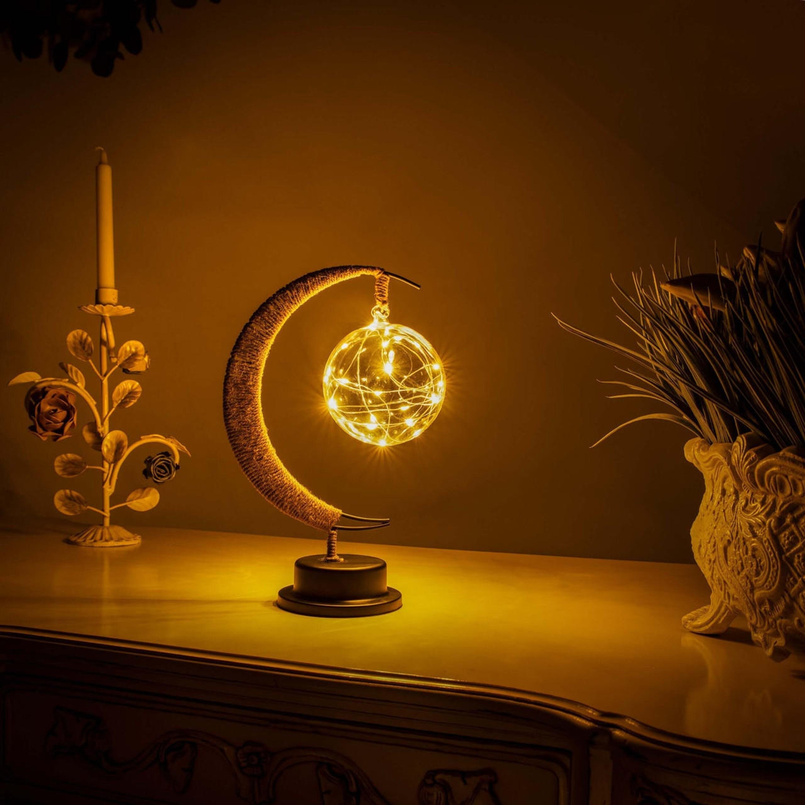 Een prachtige maanlamp met een gloeiende bol van licht, die een magische sfeer uitstraalt. De lamp staat op een zwarte basis en heeft delicate draadlampjes die een warme en betoverende ambiance creëren, perfect voor interieurdecoratie.