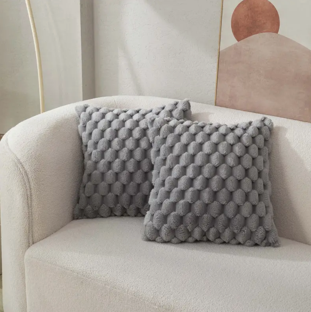 Twee grijze, donsachtige kussens met een hexagonaal patroon liggen op een comfortabele beige bank, met een minimalistisch kunstwerk op de achtergrond.