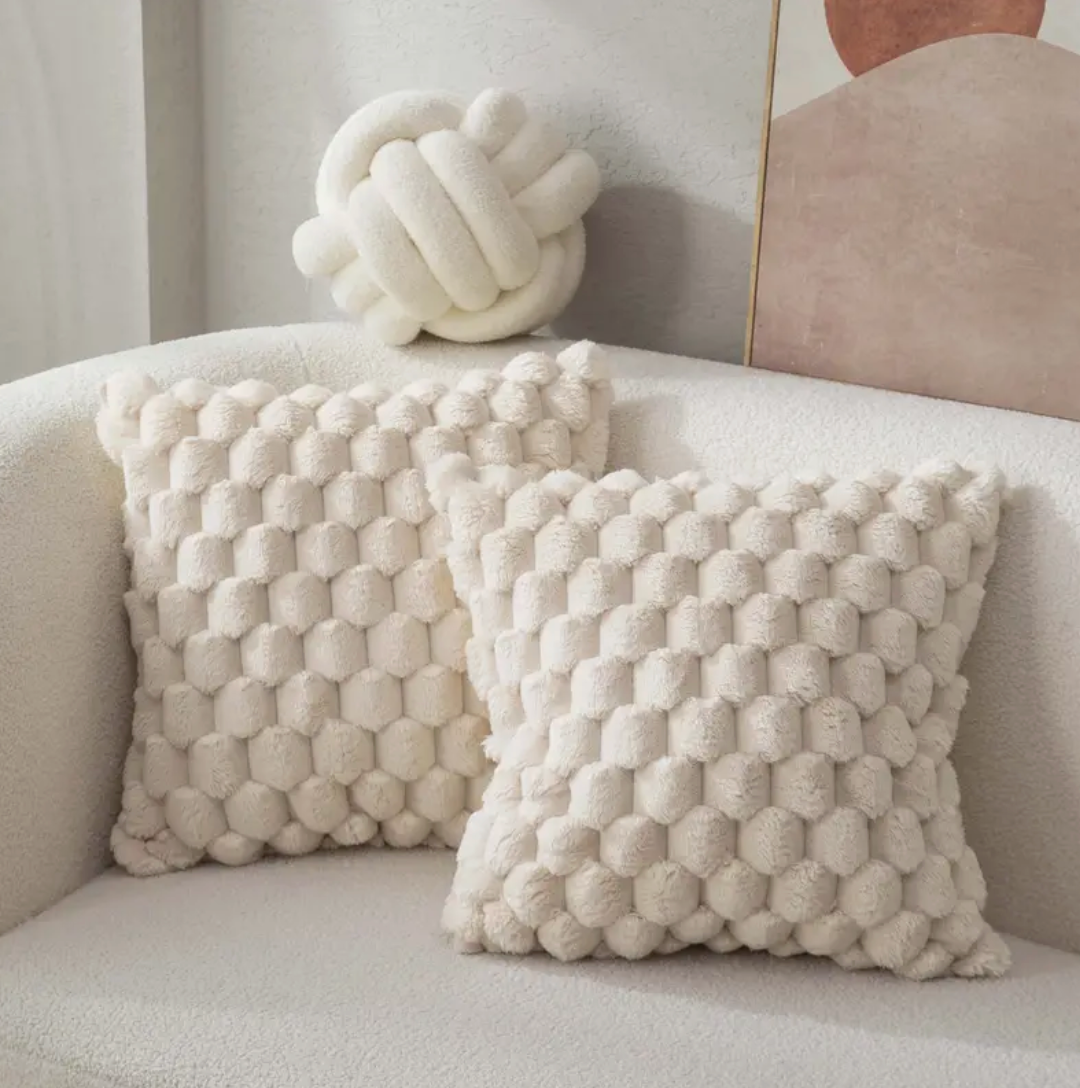 Twee donsachtige, beige kussens met een hexagonaal patroon liggen op een zachte bank, met een decoratief bolvormig kussen op de achtergrond, perfect voor een gezellige woonkamer.