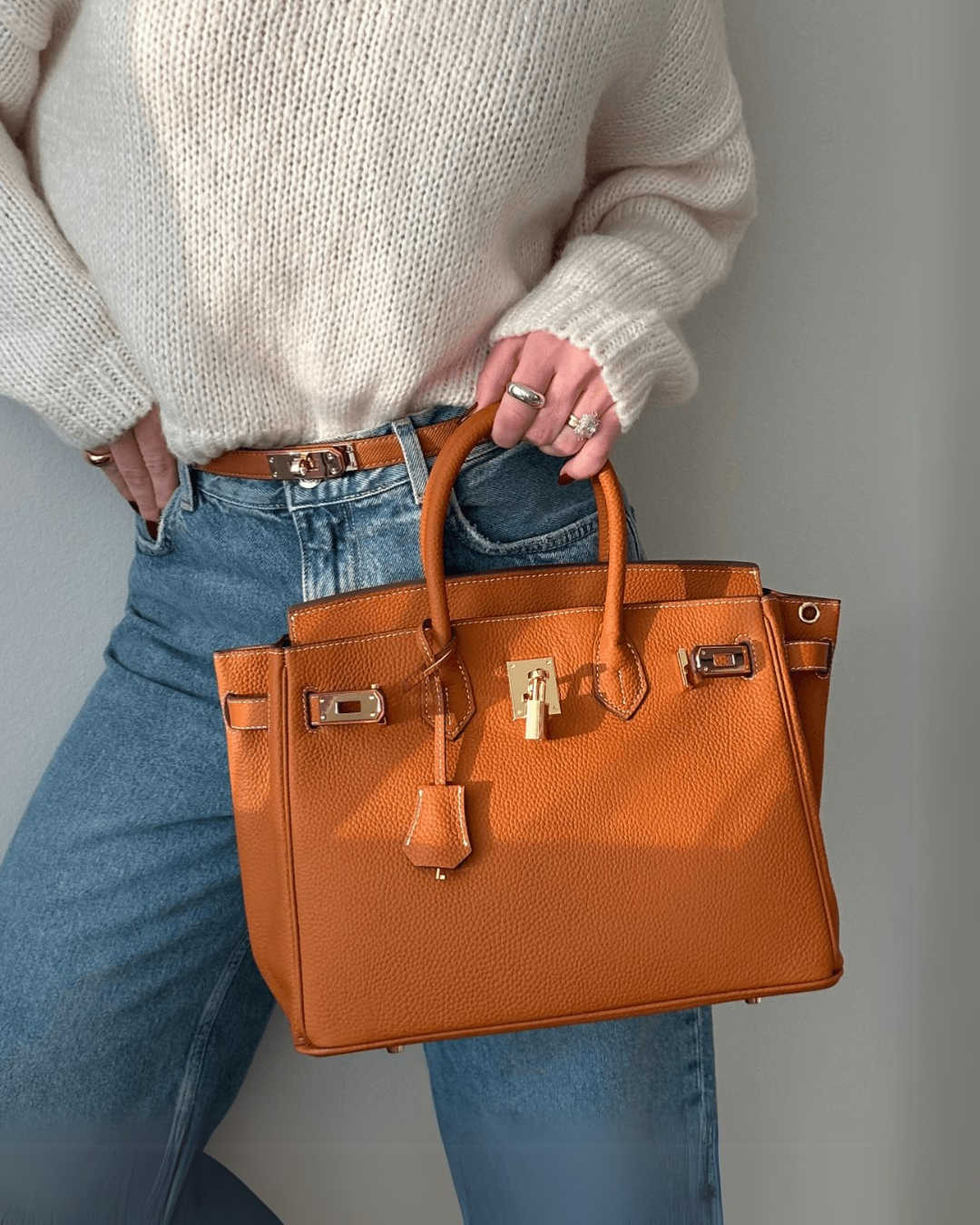 Gina tas in levendig oranje, gedragen door een persoon in een casual outfit met jeans en een crème trui. De tas straalt vrolijkheid en stijl uit, perfect voor dagelijks gebruik en bijzondere gelegenheden.