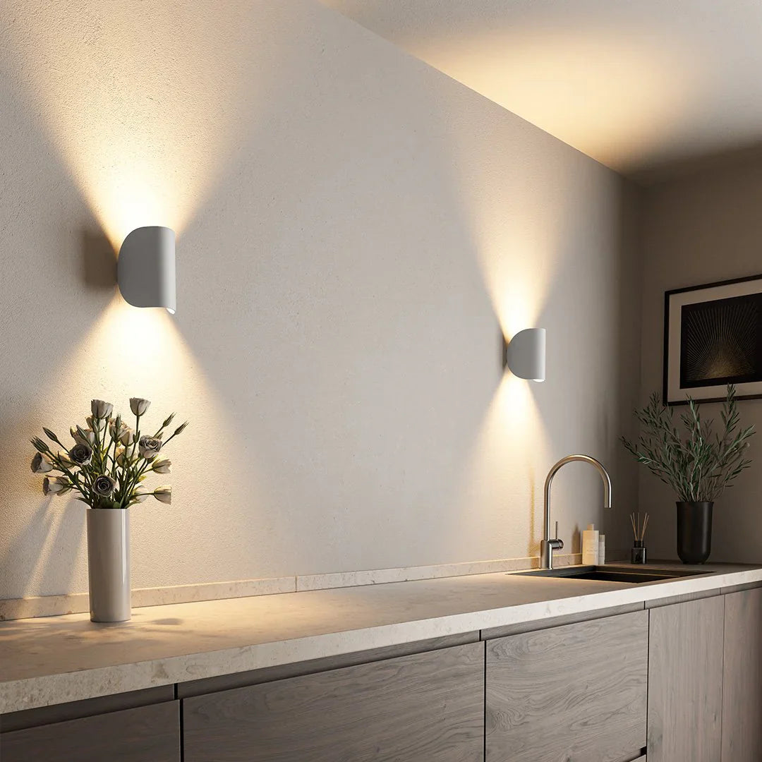 Moderne keuken met Glamora wandverlichting. Stijlvolle, minimalistische witte wandlampen stralen zacht, sfeervol licht uit, waardoor een elegante en warme sfeer ontstaat. Het strakke ontwerp aanvult de eigentijdse inrichting, waarbij een vaas met bloemen op het aanrecht en een subtiel kunstwerk aan de muur worden benadrukt.
