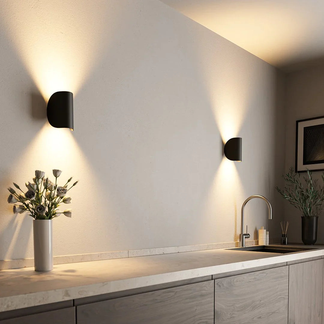 Twee stijlvolle wandlampen van Glamora verlichten zacht de muur in een moderne keuken. De lampen hebben een minimalistisch ontwerp in zwart met een verlichtingseffect dat naar boven en beneden schijnt, wat zorgt voor een elegante sfeer. Het strakke design past perfect bij het moderne interieur met een marmeren werkblad en bloemstukken.