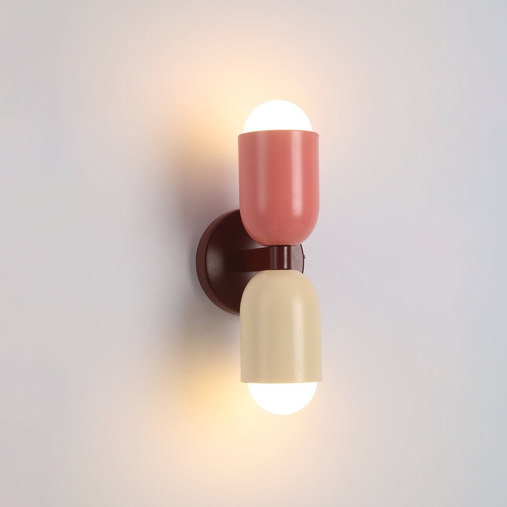 Een stijlvolle Deense design wandlamp van Glemlux met twee kleurrijke cilindrische kappen in roze en crème, gemonteerd op een ronde bruine basis, die een zacht licht uitstraalt. De moderne en minimalistische lamp is perfect om een vleugje elegantie toe te voegen aan elke interieurruimte.