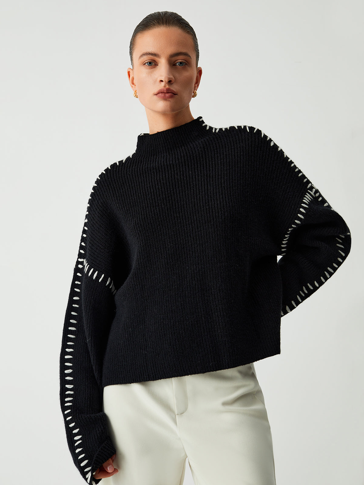 Model draagt een Helvano oversized trui in zwart met witte stikseldetails langs de schouders en mouwen. De trui heeft een hoge kraag en lange, wijde mouwen voor een comfortabele en stijlvolle uitstraling. Gecombineerd met lichte broeken voor een chique en minimalistische look.