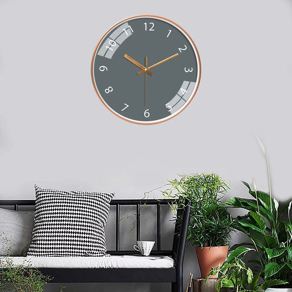 Jomera wandklok met een strak en modern design, grijze wijzerplaat en gouden wijzers. De klok hangt boven een stijlvolle bank met zwart-witte kussens en groene planten in een eigentijds ingerichte ruimte. Een elegante toevoeging aan elk interieur.