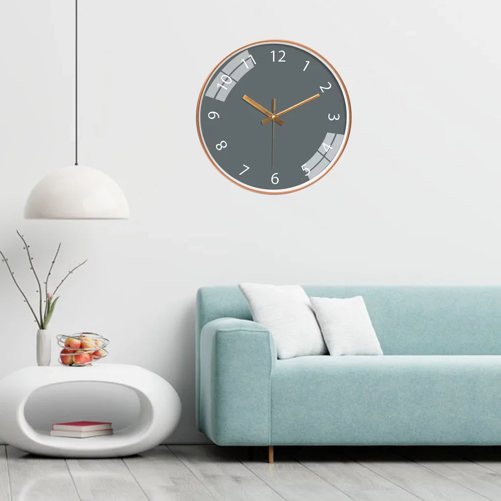 Jomera wandklok met grijze wijzerplaat en gouden wijzers, perfect passend in een modern interieur. De klok hangt boven een stijlvolle lichtblauwe bank met witte kussens. Ernaast staat een design bijzettafel met fruit en boeken, wat zorgt voor een harmonieuze en elegante uitstraling.