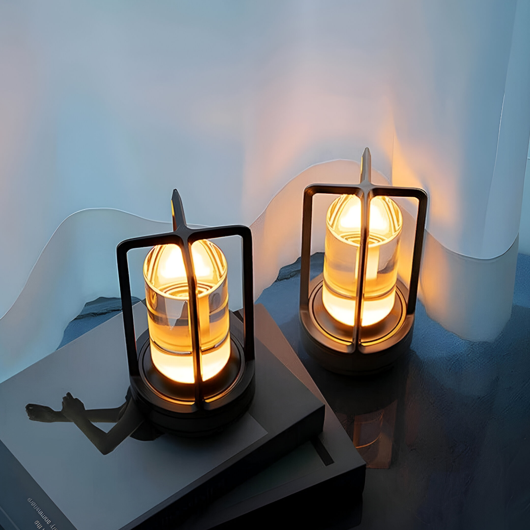  Twee stijlvolle, waterbestendige lantaarnlampen van Jovien gloeien warm op, geplaatst op een strak zwart oppervlak met een boek. Het elegante ontwerp van de lampen is voorzien van een transparant glazen hart dat is gevat in een beschermend gouden frame, waardoor een gezellige en sfeervolle sfeer in de kamer wordt gecreëerd.