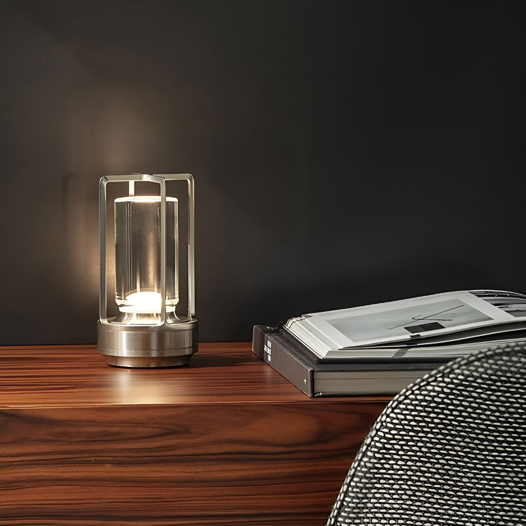 De afbeelding toont de Jovien Lamp, een stijlvolle en waterbestendige lamp met een minimalistisch ontwerp. De lamp is geplaatst op een houten bureau en straalt een warme gloed uit, wat bijdraagt aan de gezellige sfeer van de ruimte. In de achtergrond zijn een modern kantoorstoeltje en boeken te zien, wat de veelzijdigheid van de lamp in verschillende interieursettings benadrukt.
