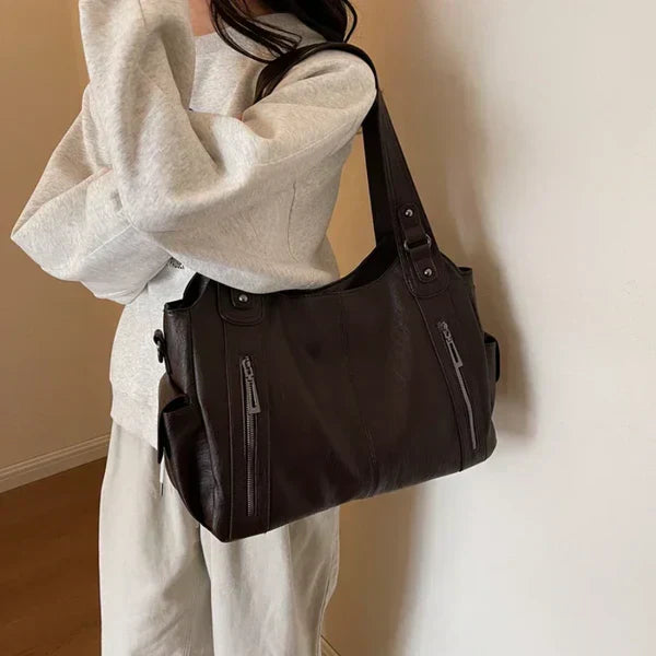 Kaldar stijlvolle leren tas in donkerbruin, gedragen door een vrouw in casual outfit. De tas heeft stevige handvatten, meerdere ritsvakken en een ruim interieur. Perfect voor dagelijks gebruik met een elegante uitstraling.