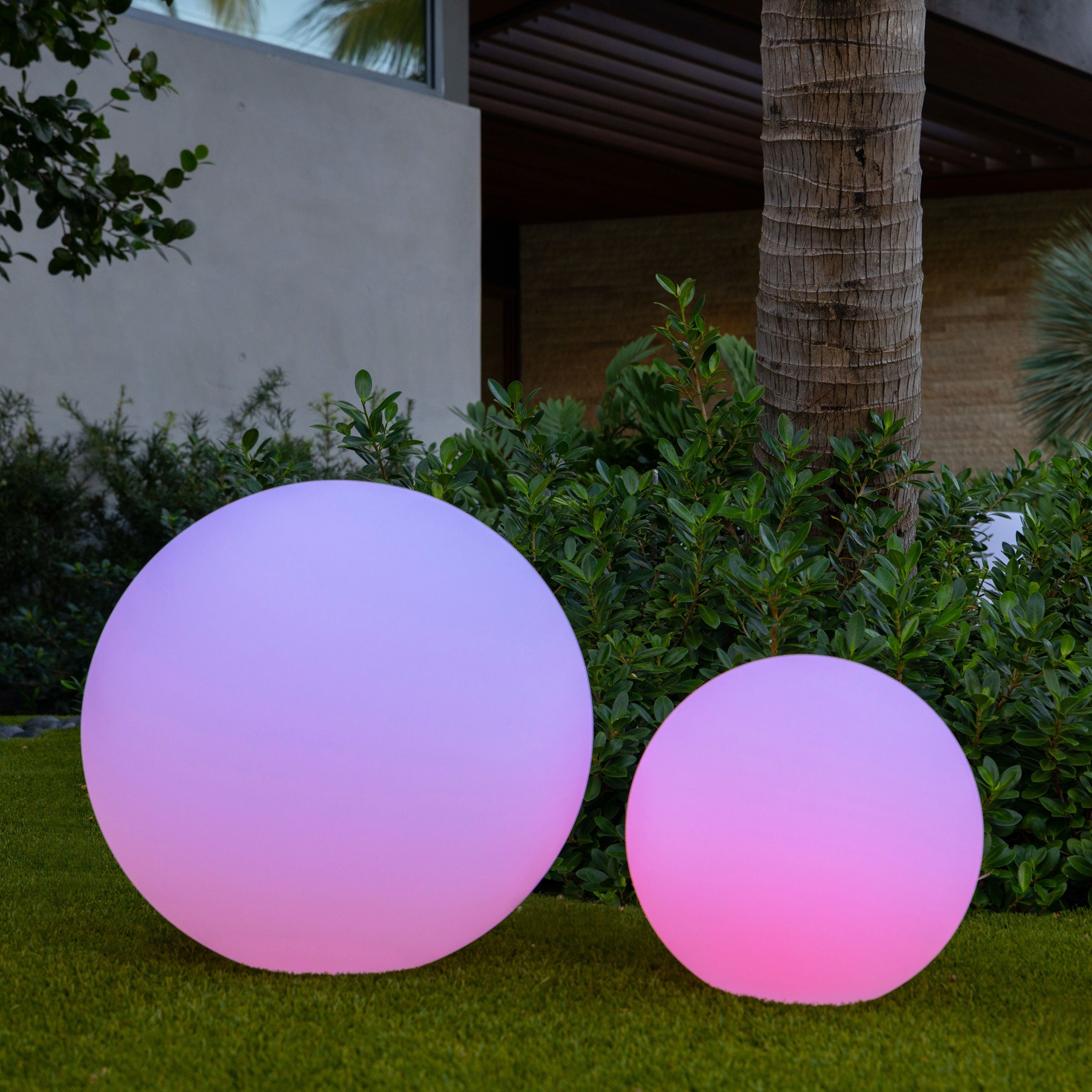 Kendra tuinverlichting met grote en kleine sferische lampen in pastelkleuren, die een tuin verlichten, met groene struiken op de achtergrond.