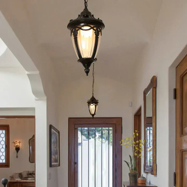 Korenia hangende lantaarn in een klassieke gang, met een elegant zwart frame en zacht warm licht. De lantaarn hangt aan een ketting en straalt stijl en charme uit, passend bij een traditioneel of landelijk interieur. De lamp is geplaatst bij de ingang en verlicht de ruimte op een sfeervolle manier.