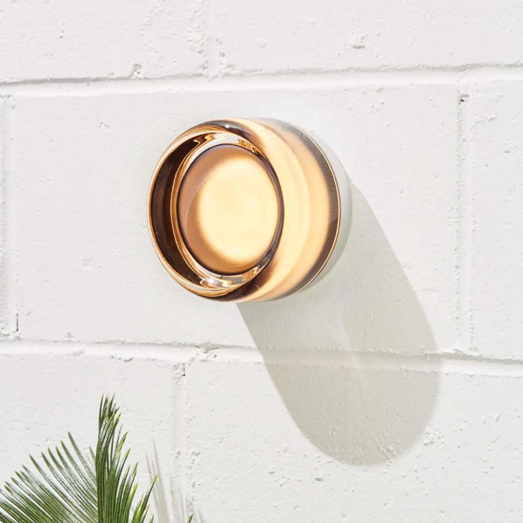 LichtAura wandlamp met een modern design, afgewerkt in koperkleur en stralend een warme gloed uit op een witte bakstenen muur.