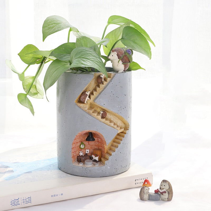 Een schattige betonnen planthouder in de vorm van een huis met een trap en egelfiguren, geplaatst op een tafel met een plant erboven. De planthouder heeft een gedetailleerde trap waar egels naar boven klimmen en een gezellig huisje met meubels erin, compleet met een lamp. Ernaast staan twee kleine egels met een paddenstoel. Het ontwerp is speels en elegant, ideaal voor een gezellige en natuurgerichte uitstraling.