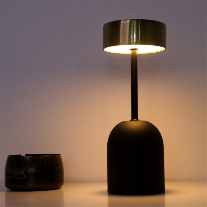 Lumeon LED-lamp met een zwarte matte voet en een metalen kap, straalt warm licht uit. De lamp heeft een modern, minimalistisch ontwerp en staat op een houten oppervlak. Ideaal voor sfeerverlichting en een stijlvolle toevoeging aan elk interieur. Energiezuinig en functioneel.
