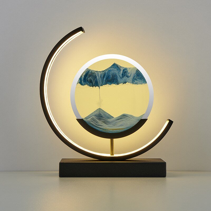 De LumiAura lamp toont een prachtig dynamisch zandlandschap in een cirkelvormig ontwerp. Het zachte, omringende licht accentueert de vloeiende beweging van het zand in blauwe tinten, die een rustgevende en betoverende sfeer creëren. Deze innovatieve lamp is een combinatie van kunst en verlichting, perfect voor moderne interieurs waar stijl en harmonie samenkomen. Het ontwerp voegt niet alleen verlichting toe, maar ook een visueel rustpunt in elke ruimte.