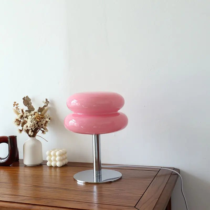 Iconische Luminore Design tafellamp in een levendige roze tint, voorzien van een dubbele glazen kap met glanzende afwerking en een elegante metalen voet. Geplaatst op een houten tafel met stijlvolle decoraties, ideaal voor een modern en eigentijds interieur
