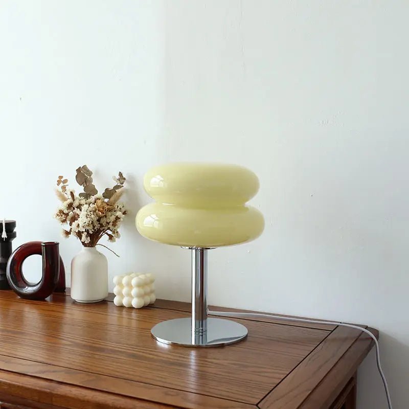 Ikonische Luminore Design tafellamp in een zachtgele tint, met een glanzende dubbele glazen kap en een slank metalen basis, geplaatst op een houten tafel naast stijlvolle decoratieve accessoires. Perfect voor een modern en verfijnd interieur.