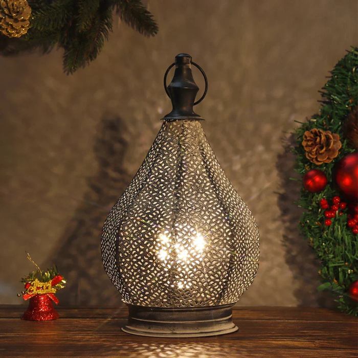 Een elegante tafel lamp met een decoratief metalen ontwerp in de vorm van een waterdruppel. Het lamplicht straalt zacht door de openingen van het fijne, metalen rooster, waardoor een sfeervolle, warme gloed ontstaat. De lamp is geplaatst op een houten tafel, omgeven door kerstdecoraties, wat een gezellige en feestelijke uitstraling geeft. Perfect voor het toevoegen van een vleugje verfijning en warmte aan elk interieur.