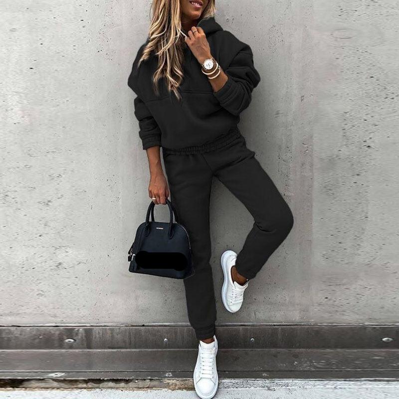 Een vrouw in een zwart joggingpak met een comfortabele trui en een broek met elastische tailleband. Ze draagt een tas en witte sneakers, met een casual, trendy uitstraling. De look is stijlvol en comfortabel, perfect voor dagelijks gebruik en ontspanning. De outfit benadrukt mode en functionaliteit, en de vrouw poseert tegen een neutrale achtergrond.