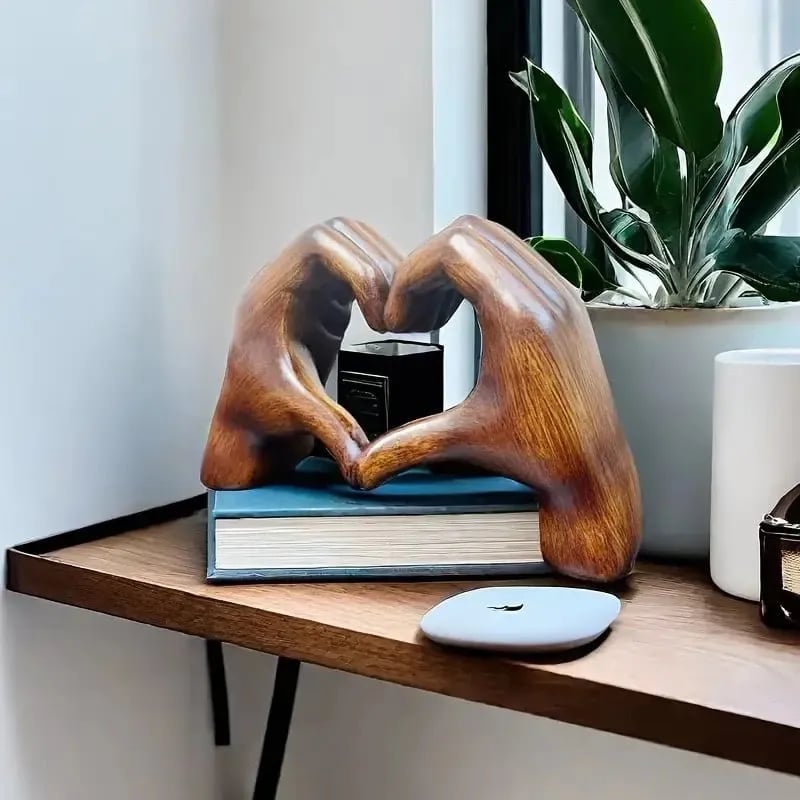 Een betoverend houten hartbeeld van Olivan, geplaatst op een boekenplank naast een plant en andere decoraties. Het kunstwerk toont twee handen die samen een hart vormen, wat zorgt voor een elegante en warme sfeer in het interieur.